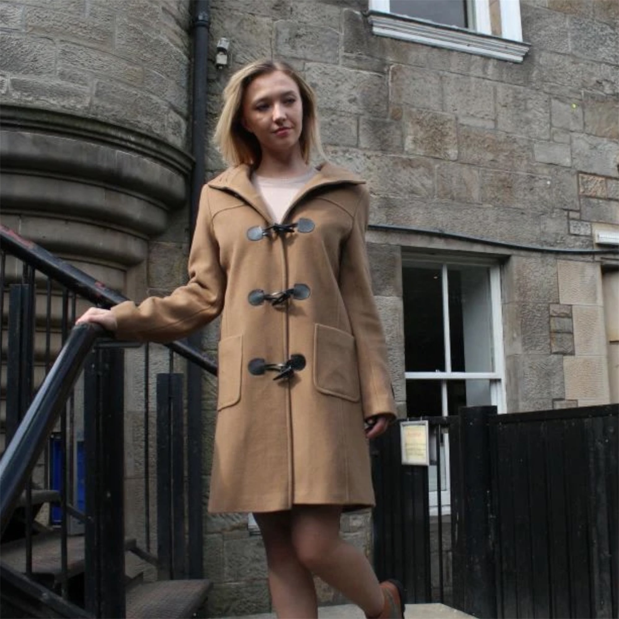 A woman standing on steps wearing a beige duffle coat with a hood, black buttons, and a visible collar. The coat has two front pockets and is paired with dark trousers.